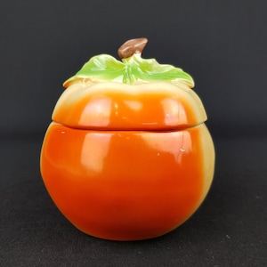 May include: A ceramic jar shaped like a peach with a lid. The lid is also shaped like a peach with a green leaf and a brown stem.