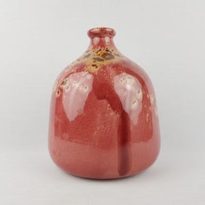 Könnte beinhalten: Eine rote Keramikvase mit rundem Körper und schmalem Hals. Die Vase hat eine glänzende Oberfläche mit einem dunkleren roten vertikalen Streifen und gesprenkelten Details in der Nähe der Oberseite. Die Vase ist etwa 20 cm hoch.