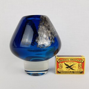 Puede incluir: Un jarr&oacute;n de vidrio azul con un dise&ntilde;o de remolino blanco y una base de vidrio transparente. El jarr&oacute;n est&aacute; sobre una superficie blanca junto a una caja de cerillas.