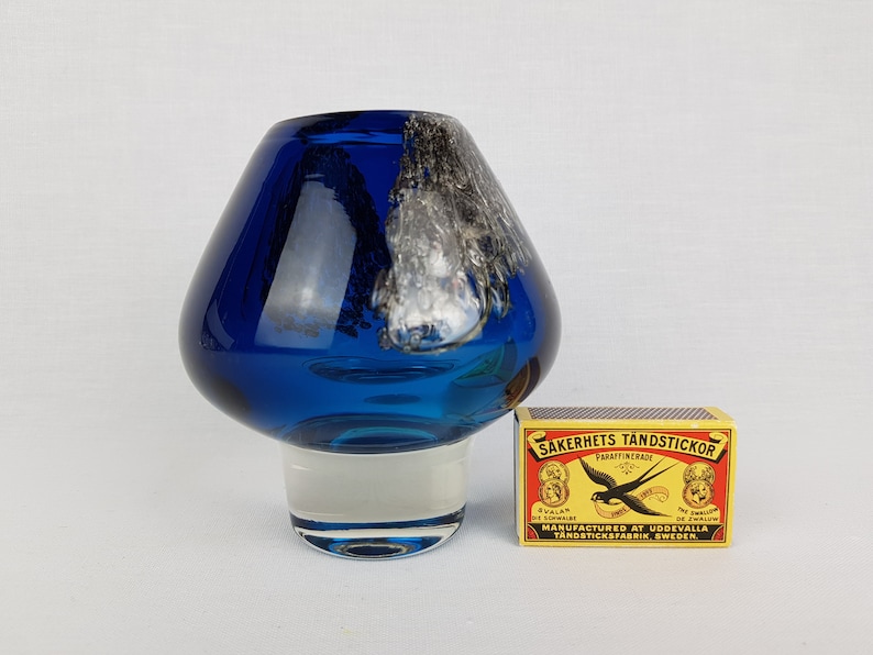 Puede incluir: Un jarr&oacute;n de vidrio azul con un dise&ntilde;o de remolino blanco y una base de vidrio transparente. El jarr&oacute;n est&aacute; sobre una superficie blanca junto a una caja de cerillas.