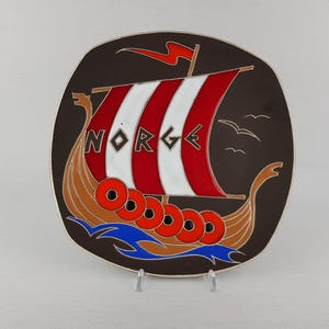 May include: A decorative plate with a Viking ship design. The ship has a red and white striped sail, with the word "NORGE" on the sail. The ship is brown with red circles and blue waves. The plate is dark brown with a white background.