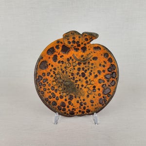 Puede incluir: Un plato de cerámica redondo con forma de manzana y un tallo en la parte superior. La superficie presenta un esmalte naranja vibrante con una textura de color marrón oscuro, moteada y con cráteres. El plato se exhibe sobre un soporte de plástico transparente.