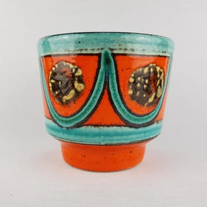 Op de afbeelding: Een keramische plantenbak met een levendig ontwerp. De plantenbak heeft een feloranje basis en body, geaccentueerd met turquoise banden aan de rand en basis. De oranje body is versierd met donkergroene en zwarte gebogen lijnen en cirkelvormige patronen met gele en bruine accenten.