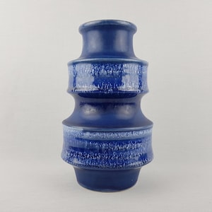 Puede incluir: Un jarrón de cerámica azul con un diseño texturizado y en capas. El jarrón tiene forma cilíndrica con una base ancha y una parte superior más estrecha. El azul tiene un efecto degradado, con tonos más claros en las zonas texturizadas.
