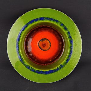 Puede incluir: Un plato de cerámica con un diseño vibrante. El plato presenta un borde exterior verde brillante, una banda moteada azul y un diseño central de círculos concéntricos en tonos naranja y marrón. El plato tiene un acabado brillante.