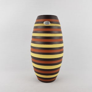 May include: A tall, oval-shaped vase with horizontal stripes in alternating colors of brown, yellow, and orange. The vase has a dark brown top and a small, round sticker near the top. The design is symmetrical and the vase is set against a plain white background.