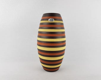 May include: A tall, oval-shaped vase with horizontal stripes in alternating colors of brown, yellow, and orange. The vase has a dark brown top and a small, round sticker near the top. The design is symmetrical and the vase is set against a plain white background.
