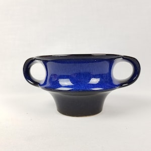 Könnte beinhalten: Eine blaue und schwarze Keramiktasse mit zwei Henkeln. Die Tasse hat einen abgerundeten Boden und einen ausgestellten Rand.