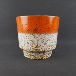 Op de afbeelding: Een keramische plantenpot met een tweekleurig ontwerp, bestaande uit een glanzend oranje bovendeel en een wit onderste deel. Het gehele oppervlak is voorzien van een bruine spikkeltextuur die geconcentreerder is op de witte basis en rand.