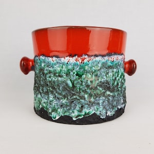 Op de afbeelding: Een rode, groene en zwarte keramische plantenpot met twee handvatten. De pot heeft een ruw, getextureerd oppervlak.