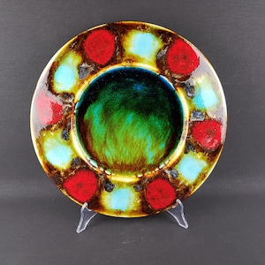 May include: A decorative ceramic plate with a vibrant design. The plate features a central green area surrounded by a ring of red, blue, and yellow polka dots. The plate is displayed on a clear stand, showcasing its colorful and artistic appeal.