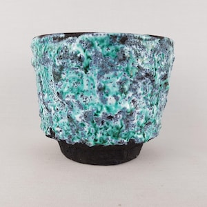 May include: A ceramic cup featuring a dark, matte black base and a heavily textured, cratered upper section. The surface is glazed in a vibrant mix of turquoise, emerald green, and grey tones, creating a rugged, uneven appearance with deep crevices.