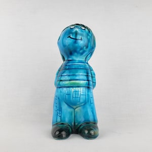 Könnte beinhalten: Eine Keramikfigur einer stehenden Person mit den Händen hinter dem Rücken, die eine glänzende blaue Glasur mit dunkelblauen Akzenten aufweist. Die Figur trägt ein gestreiftes Oberteil und eine Hose, hat ein lächelndes Gesicht und dunkle Schuhe.