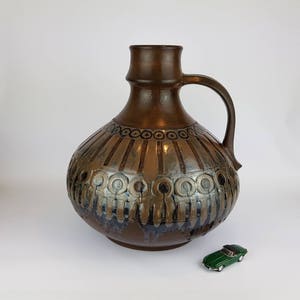 May include: A large, brown ceramic vase with a wide, round body and a narrow neck. The vase has a handle and is decorated with a geometric pattern of circles and lines in shades of brown, black, and silver.