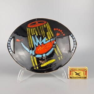 May include: Black ceramic serving dish with a colorful abstract design. The dish features a black background with yellow, blue, and orange geometric shapes. It has two handles and is displayed on a clear stand.