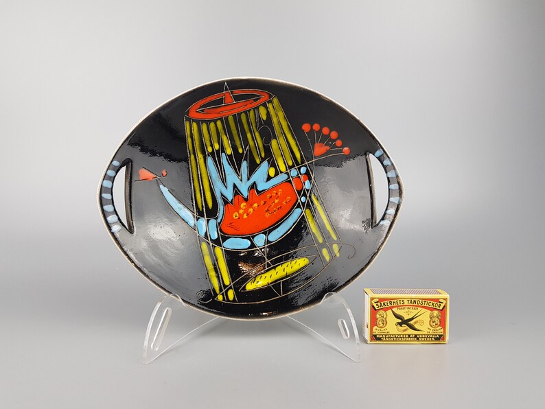 May include: Black ceramic serving dish with a colorful abstract design. The dish features a black background with yellow, blue, and orange geometric shapes. It has two handles and is displayed on a clear stand.