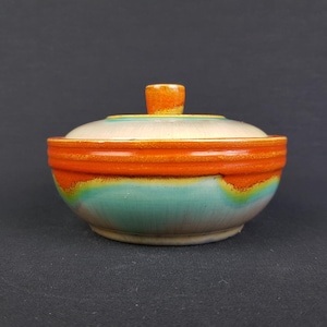 Puede incluir: Un cuenco de cerámica con tapa, con un esmalte azul, verde, amarillo y naranja. El cuenco tiene una forma redondeada y un borde ancho.