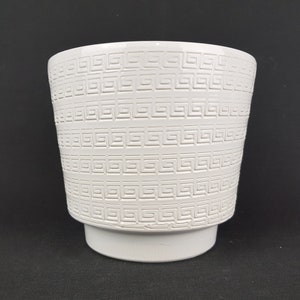 May include: A white ceramic planter featuring a repeating embossed Greek key pattern around the exterior. The vessel has a wide, open top that tapers down to a smooth, solid base, providing a clean and structured appearance for this container.