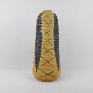 Może przedstawiać: Wysoki, ceramiczny wazon z teksturowaną, beżową podstawą i wąską szyjką. Wazon jest ozdobiony pionowymi niebieskimi paskami i czarnym wzorem krzyżykowym. Wazon ma około 25 cm wysokości.