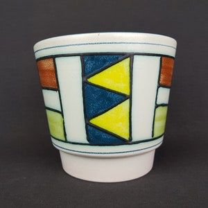 Op de afbeelding: Een witte keramische plantenpot met een geometrisch ontwerp in tinten blauw, geel, groen en bruin. De pot heeft een brede basis en een licht uitlopende bovenkant.