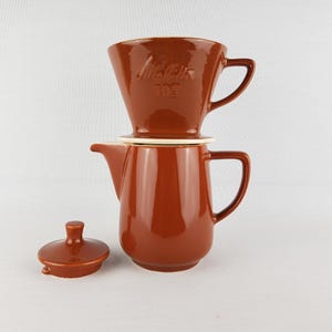May include: A glossy brown ceramic coffee pot with a matching lid placed to its side and a Melitta 102 coffee filter cone stacked on top. The coffee pot features a side handle and a spout, while the filter cone has the embossed text Melitta 102.