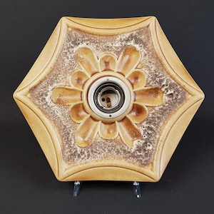 Op de afbeelding: Een zeshoekige keramische plafondlamp met een beige en bruine geglazuurde afwerking en een centraal bloemachtig reliëfpatroon. Het midden bevat een ronde metalen fitting voor een gloeilamp. Het oppervlak heeft een textuur met vlekken.
