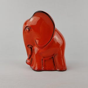 May include: A ceramic elephant figurine in a vibrant orange hue with black outlining. The elephant has large ears, a trunk, and detailed facial features. The figurine is a decorative item, likely for a shelf or desk.