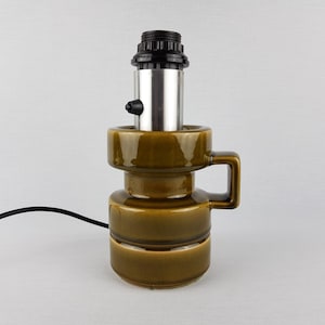 Op de afbeelding: Een bruine keramische tafellamp met een zilveren metalen kap en een zwart snoer. De lamp heeft een uniek ontwerp met een handvat aan de zijkant.