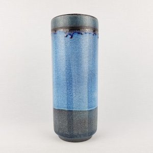 May include: A tall, cylindrical ceramic vase with a two-tone design. The upper section is light blue with vertical stripes, while the bottom is a darker grey. The rim is a darker blue-grey. The vase has a glossy finish.