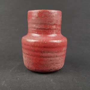 Op de afbeelding: Een kleine, cilindrische keramische vaas met een textuurrijke, gespikkelde rode glazuurlaag. De vaas heeft een brede, ronde basis die taps toeloopt naar een smallere hals, met horizontale ribbels rondom het lichaam en een gladde, open bovenrand.
