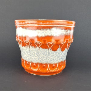 Puede incluir: Un macetero vintage de cerámica naranja y blanco con un diseño decorativo. El macetero tiene un borde ancho y una base redondeada.