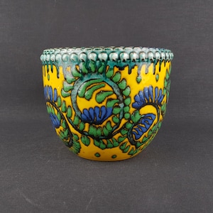 Puede incluir: Un cuenco de cerámica con una base de color amarillo brillante, que presenta un patrón repetitivo de motivos en forma de hoja en azul oscuro y verde. El borde superior está decorado con una textura de cuentas en relieve en tono turquesa.