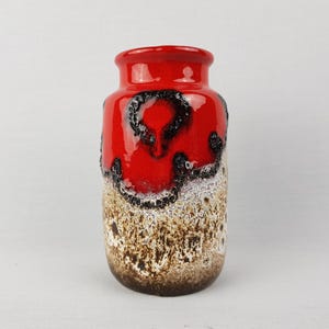 Puede incluir: Un jarrón de cerámica rojo vibrante con un diseño texturizado y moteado. La parte superior y el interior son de color rojo brillante, mientras que el cuerpo presenta una mezcla de texturas negras, blancas y marrones. El jarrón tiene forma cilíndrica.