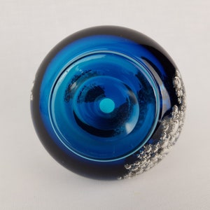 Puede incluir: Un pisapapeles de vidrio azul con un dise&ntilde;o plateado de burbujas en el lateral. El pisapapeles tiene un centro transparente con un anillo azul alrededor.