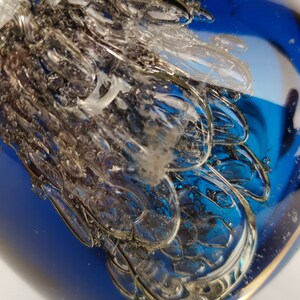 Puede incluir: Primer plano de un pisapapeles de vidrio azul con un dise&ntilde;o claro y texturizado en su interior. El vidrio es transparente y tiene una superficie lisa y brillante.