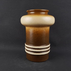 Può includere: Un vaso cilindrico in ceramica con una combinazione di marrone e beige. Il vaso presenta una parte superiore arrotondata, una fascia beige più chiara e tre strisce orizzontali bianche vicino alla base. Un oggetto decorativo.