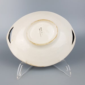 May include: White ceramic serving dish with a raised center and two black handles. The dish has a slightly irregular shape and is displayed on a clear acrylic stand. The words "MADE IN ITALY" are printed on the bottom.