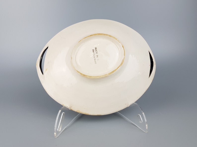 May include: White ceramic serving dish with a raised center and two black handles. The dish has a slightly irregular shape and is displayed on a clear acrylic stand. The words "MADE IN ITALY" are printed on the bottom.