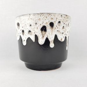 Puede incluir: Jardinera de cerámica en blanco y negro con un diseño único de esmalte goteado. La parte superior presenta un esmalte blanco texturizado con manchas marrones oscuras, mientras que la sección inferior es negra. La jardinera tiene forma cilíndrica con una base ligeramente ensanchada.