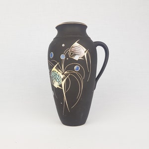 May include: Black ceramic vase with a gold and blue fish design. The vase has a handle and a rounded body.