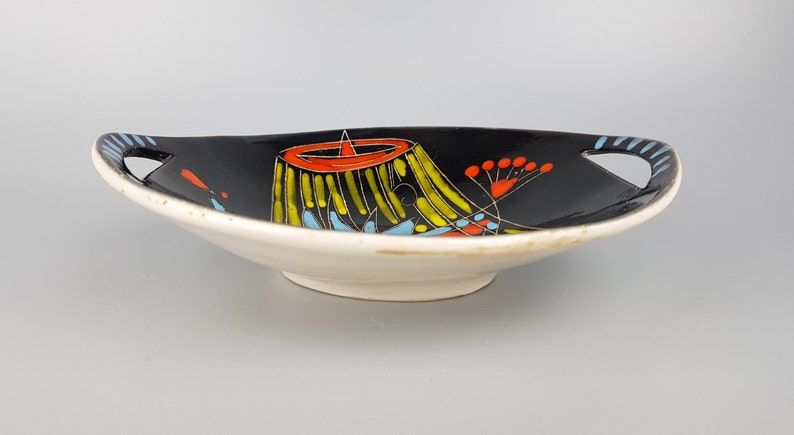 May include: A ceramic serving dish with a black interior and a white exterior. The dish features a colorful abstract design with yellow, red, and blue accents. It has handles with blue stripes and measures approximately 12 inches in length.