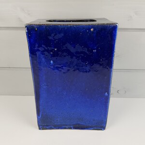 Puede incluir: Un jarrón cuadrado de cerámica con un esmalte azul oscuro. El jarrón tiene una superficie ligeramente texturizada y un borde marrón oscuro.