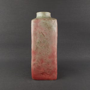 May include: A rectangular ceramic vase with a narrow neck. The vase has a gradient color scheme, transitioning from a reddish-pink at the base to a light green at the top. The surface has a textured, speckled appearance.