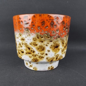 Pode incluir: Um vaso de cerâmica com um design salpicado. A seção superior é laranja, passando para branco e marrom no meio, e uma base branca. O vaso tem formato cilíndrico e é adequado para plantas de interior.