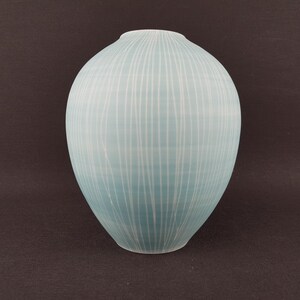 Puede incluir: Un jarrón de cerámica azul claro con forma redondeada y cuello estrecho. El jarrón está decorado con líneas blancas verticales. El jarrón mide aproximadamente 25 cm de altura.