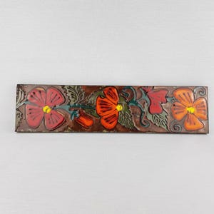 Può includere: Una piastrella di ceramica rettangolare con sfondo marrone e un motivo floreale. I fiori sono rossi, arancioni e gialli con foglie e steli verdi.