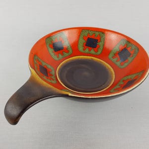 Op de afbeelding: Een keramische serveerschaal met een handvat. De schaal is voornamelijk oranje met een donkerbruin interieur en handvat. Het heeft een decoratief patroon van groene en zwarte vierkanten. De schaal is ongeveer 20 cm lang.