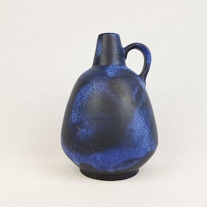 May include: A ceramic pitcher with a rounded body and a single handle. The surface features a mottled glaze with deep black and vibrant blue colors, creating a textured appearance. The neck is narrow and tapers slightly toward the top opening.