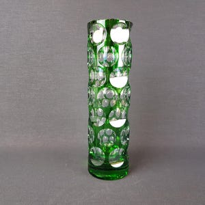 Puede incluir: Un jarrón alto y cilíndrico de vidrio verde con un patrón repetitivo de grandes recortes circulares y claros. El jarrón mide aproximadamente 20 cm de alto y tiene un acabado pulido que refleja la luz. El jarrón está vacío y sobre una superficie gris.