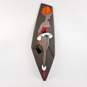 May include: A decorative wall plaque depicting a woman in a red top, white skirt, and fishnet stockings. The figure has orange hair and holds a black top hat. The background is a dark brown, textured shape.